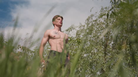 blue sky with trees swaying around, tourist stands amid riverside grasses loosening band, sunlit torso showing muscles and tattoos, leafy branches framing scene, calm relief after steep wooded climb
