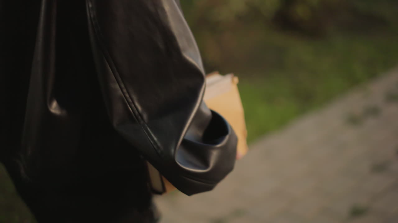 Pessoa lendo ao ar livre à noite, indivíduo praticando atividade literária noturna ao ar livre, pessoa vista ao ar livre em um parque da cidade durante a noite segurando um livro perto de uma bolsa e um acessório de couro