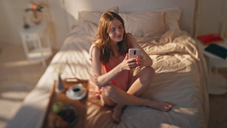 happy teenager holding smartphone resting in sunny bedroom. dreamy model enjoy