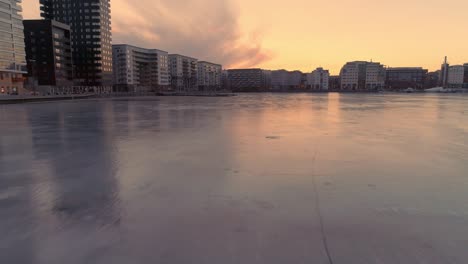 drone shot flying over ice, frozen bay and up over modern apartment buildings in stockholm, sweden. aerial view of contemporary residential district at sunset