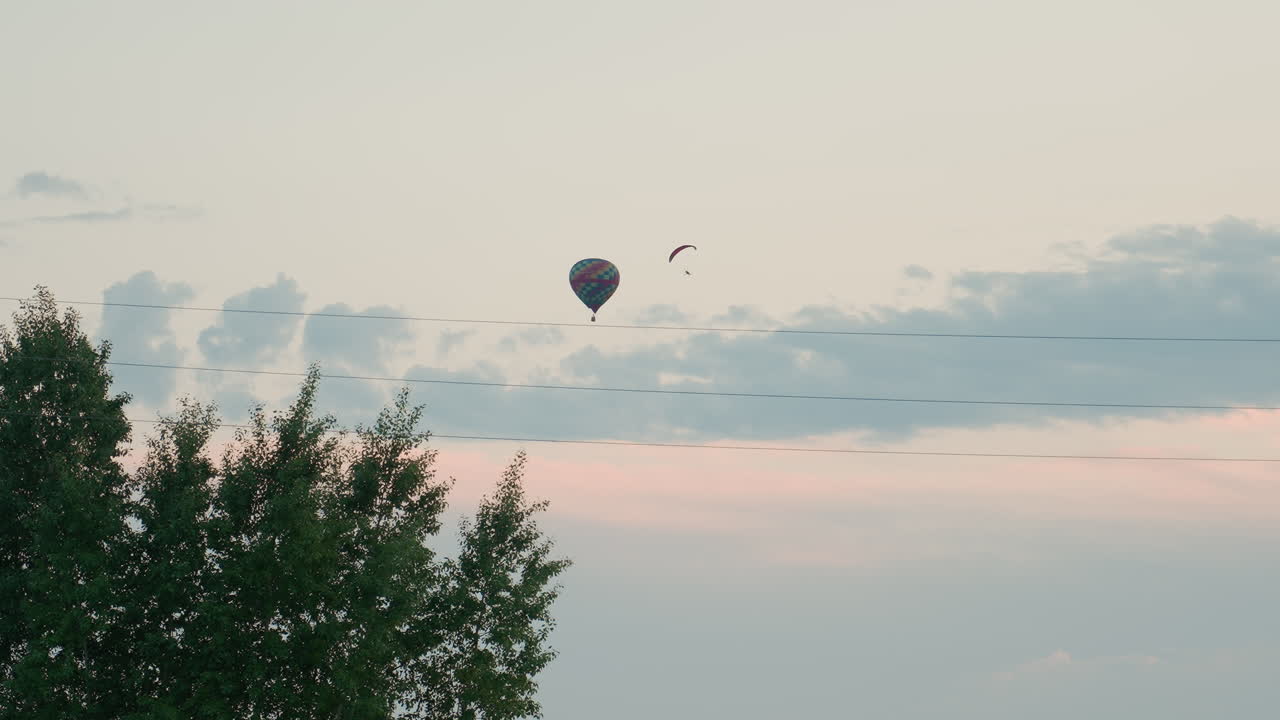 distant figures of paraglider pilot and multicolored hot air balloon drifting across pastel dusk sky above treetops and power lines conveying serene aerial adventure gentle breeze soft clouds