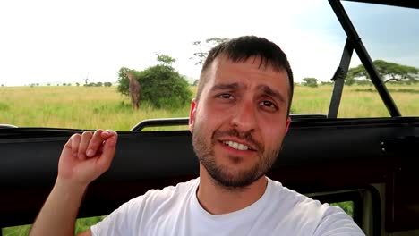 man looking into the camera explaining with hand gestures about the giraffe in the background on a safari
