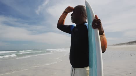 hombre mayor afroamericano de pie en una playa sosteniendo una tabla de surf y mirando hacia el mar