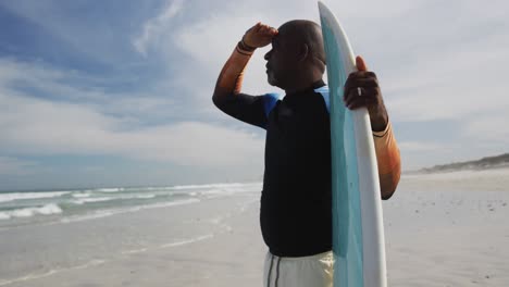 african american senior man standing on a beach holding surfboard and looking out to sea