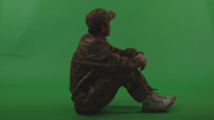 Full Body Of Asian Male Soldier Looking Around Then Touching His Head Being Sad Due To The War While Sitting On The Floor On Green Screen Background
