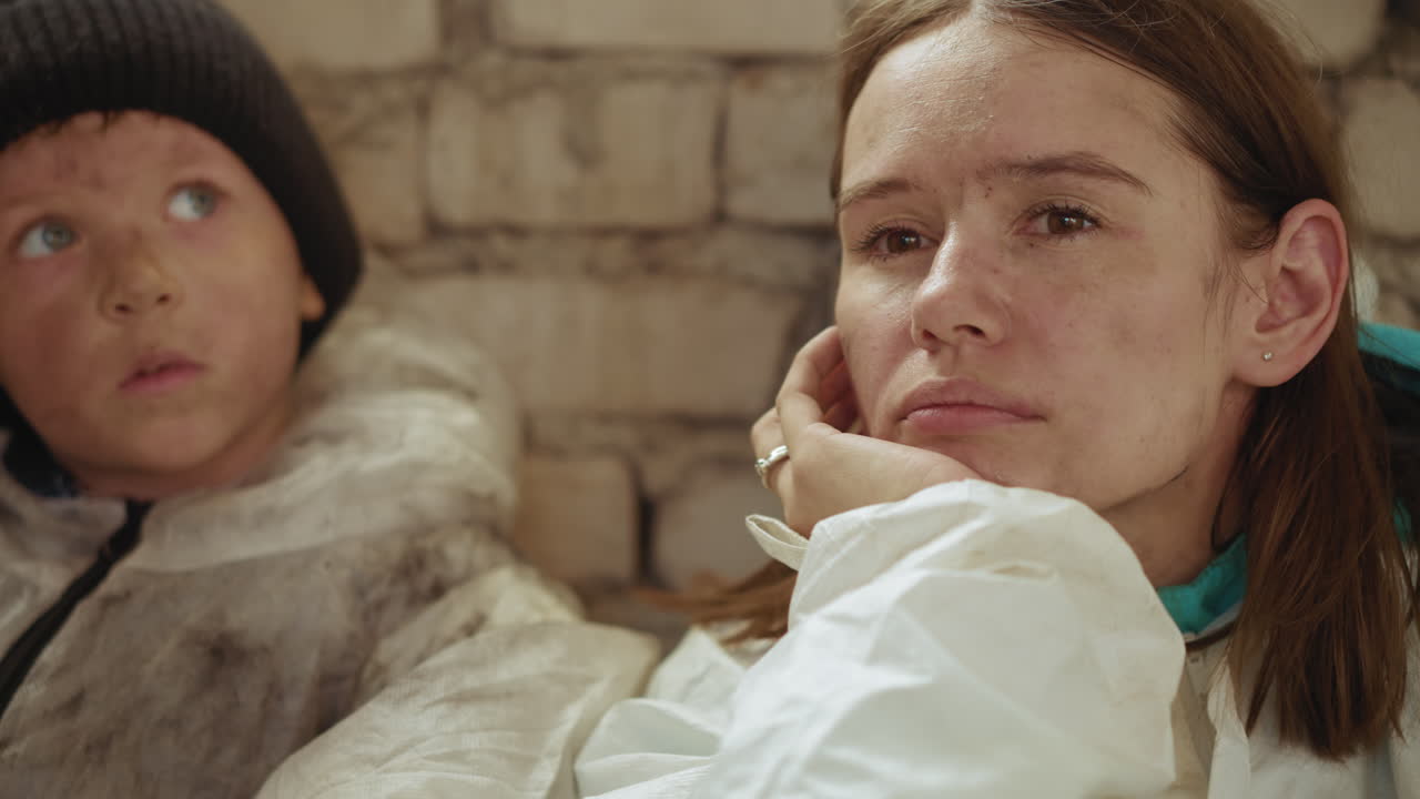 Close up of exhausted woman and dirty faced boy sitting against brick wall wearing worn clothing, gazing in silence with solemn expressions, reflecting deep thought and hardship