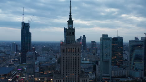 Elevated-slide-and-pan-shot-of-Palace-of-Culture-and-Science-at-dusk.-Old-high-rise-building-with-spire-and-tower-clock.-modern-skyscrapers-in-background.-Warsaw,-Poland