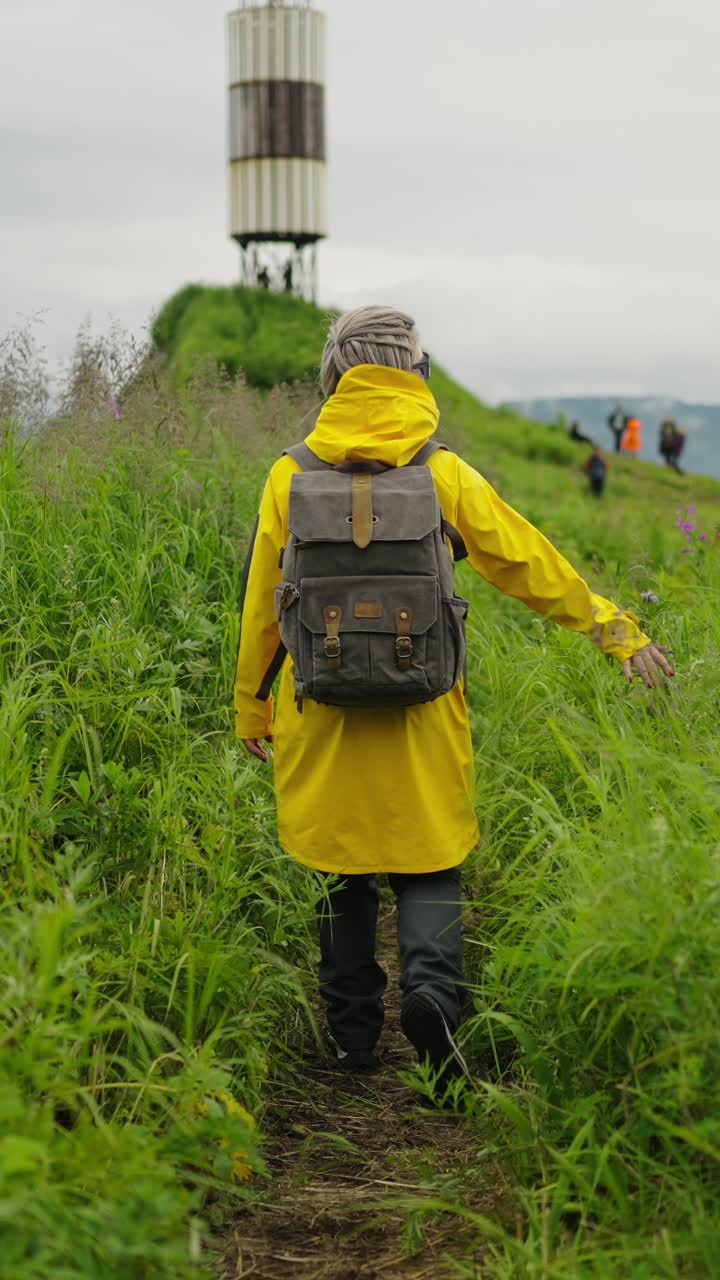 Donna con impermeabile giallo che fa trekking su una collina erbosa verso un faro.
