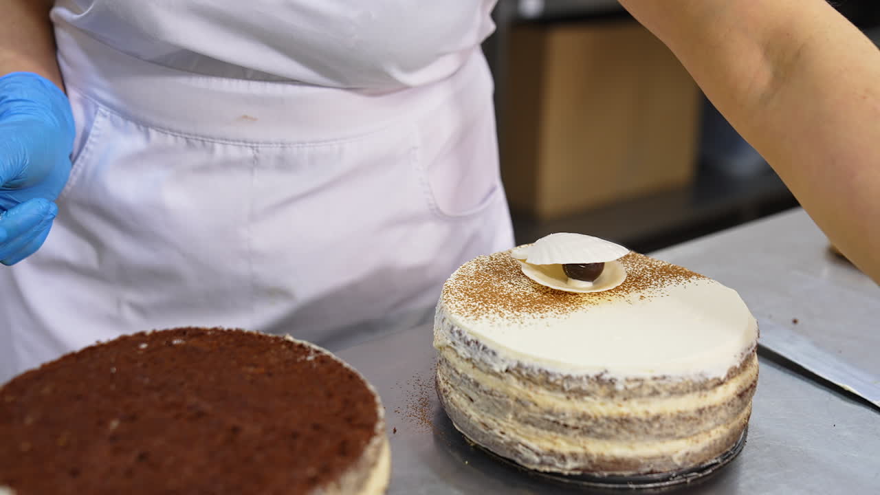 Confectioner's hands decorating the cake. Gloved hand puts white chocolate shell on top of the cake and black chocolate beads.