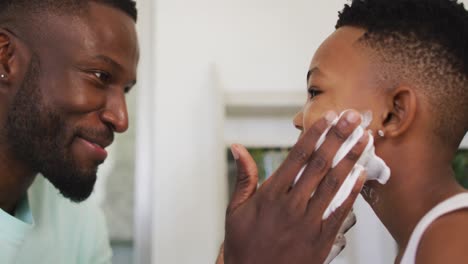 padre afroamericano poniendo crema de afeitar en la boca de su hijo y riendo juntos