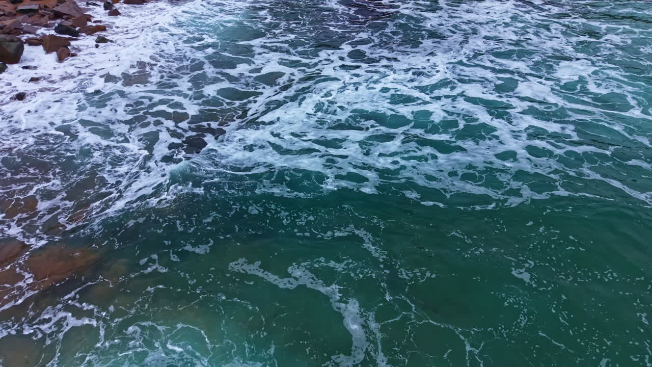 As ondas quebram em uma costa rochosa durante o dia, com espuma branca se formando na superfície da água.