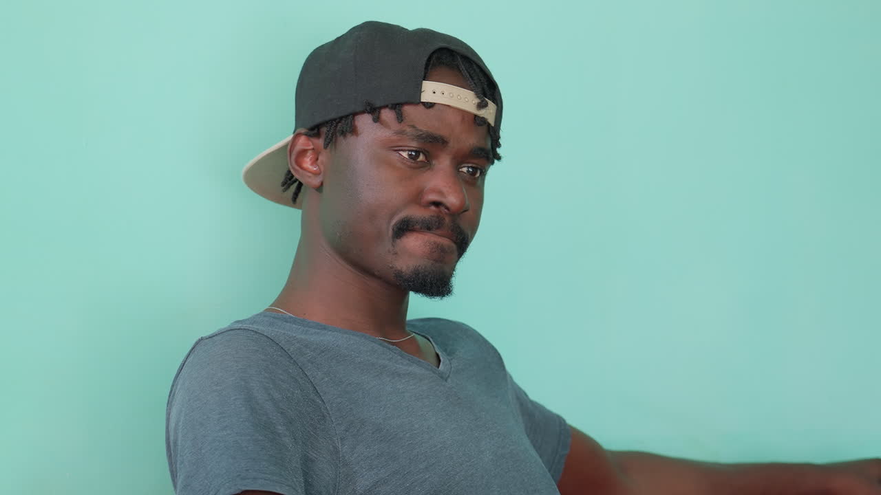 Close up of dark skin man in backward cap turning face sideways with serious thoughtful expression while sitting against mint green wall in casual streetwear