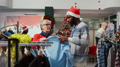 Cliente-Anciano-Navegando-Por-La-Ropa-En-La-Sección-De-Hombres-De-La-Tienda-De-Moda-Del-Centro-Comercial-Ornamentado-De-Navidad-Festiva,-Siendo-Asistido-Por-Un-Empleado-Para-Encontrar-Una-Camisa-Para-Regalarle-A-Su-Esposo-Para-La-Próxima-Temporada-De-Vacaciones-De-Invierno