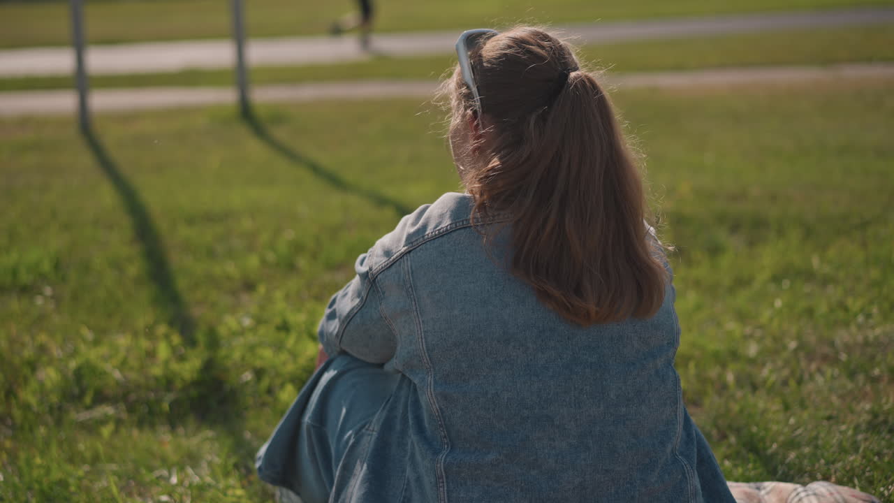 Kaukasisk kvinde sidder i græsset og vinker under et telefonopkald, varmt sollys og lange skygger, venlig hilsen til forbipasserende, influencer-stil gestik og afslappet social snak, afslappet picnicstemning.