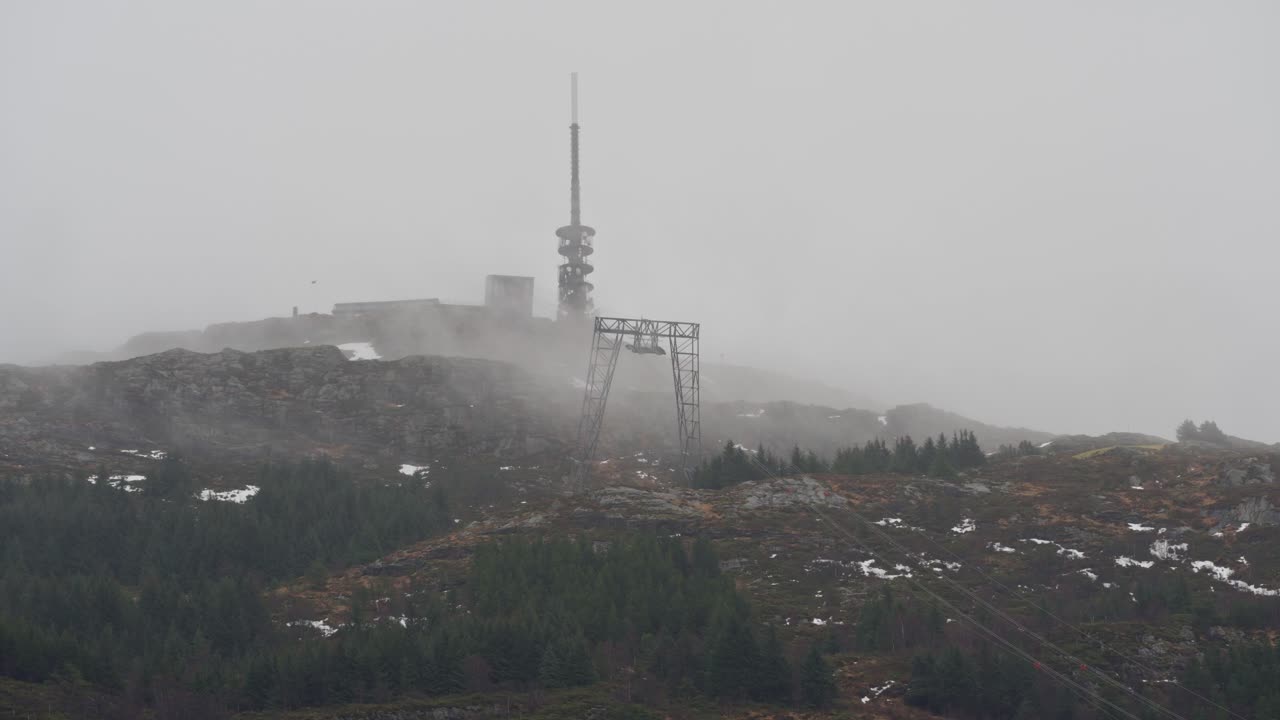 Mast on top of Ulriken mountain in Bergen, Norway, surrounded by thick fog and rain. Gondola wires stretch across the scene as clouds move past.