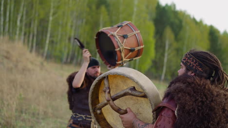 Man-in-folk-clothes-taps-drum-with-friend-against-forest