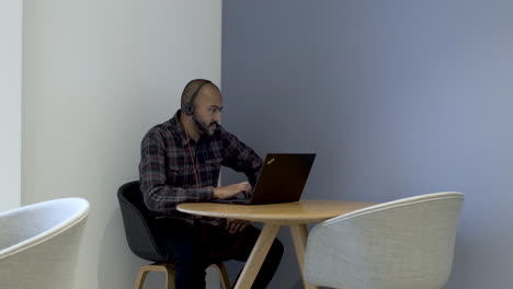 un empleado de oficina asiático que trabaja en su computadora portátil en una mesa pequeña, el hombre que usa un auricular con micrófono y navega a través de diferentes programas usando el panel táctil