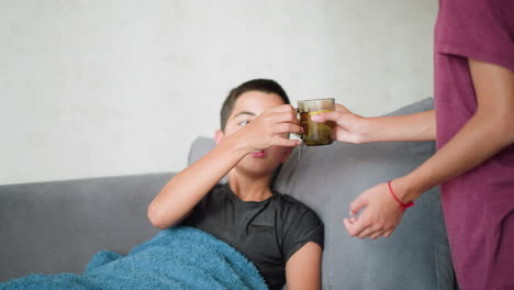 sick boy resting on couch collects cup of tea from sister and drinks it for comfort, with red lace bracelet visible on her wrist as she offers the drink