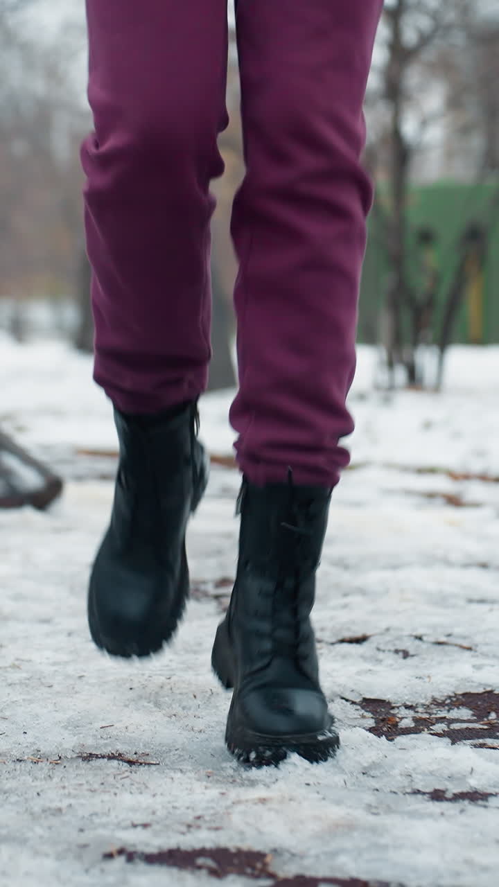 vista de la pierna de una persona en traje de invierno corriendo debido al frío en un parque al aire libre con nieve, entorno urbano con patio de recreo en el fondo, botas visibles, entrenamiento en clima frío