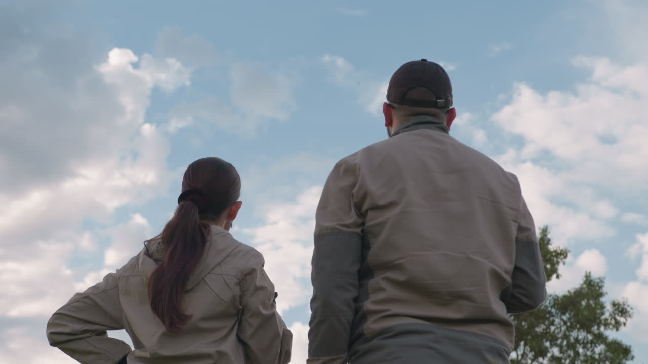 Man in black cap stands beside woman as both gaze intently into distance, sky filled with soft scattered clouds and bright blue patches above them