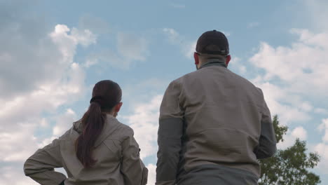 man in black cap stands beside woman as both gaze intently into distance, sky filled with soft scattered clouds and bright blue patches above them