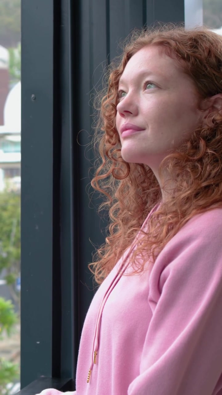 Vertical video: Adjusting hoodie woman in pink resting on balcony railing lifting gaze with pendant