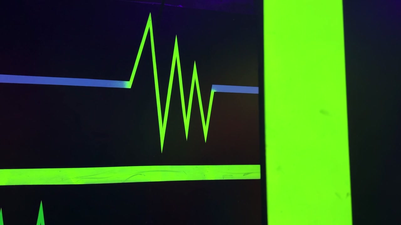 Un primo piano di un impulso cardiaco verde neon luminoso dipinto su una parete scura. La vivace grafica ultravioletta presenta accenti blu e rossi, illuminata da luce nera all'interno di un centro di intrattenimento