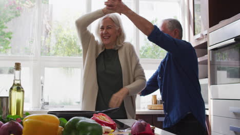 happy senior caucasian couple at home dancing in the kitchen and smiling while preparing a meal