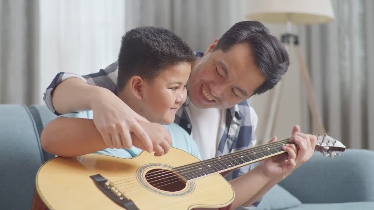 アジア人の父親が息子にソファでギターを弾くことを教えています