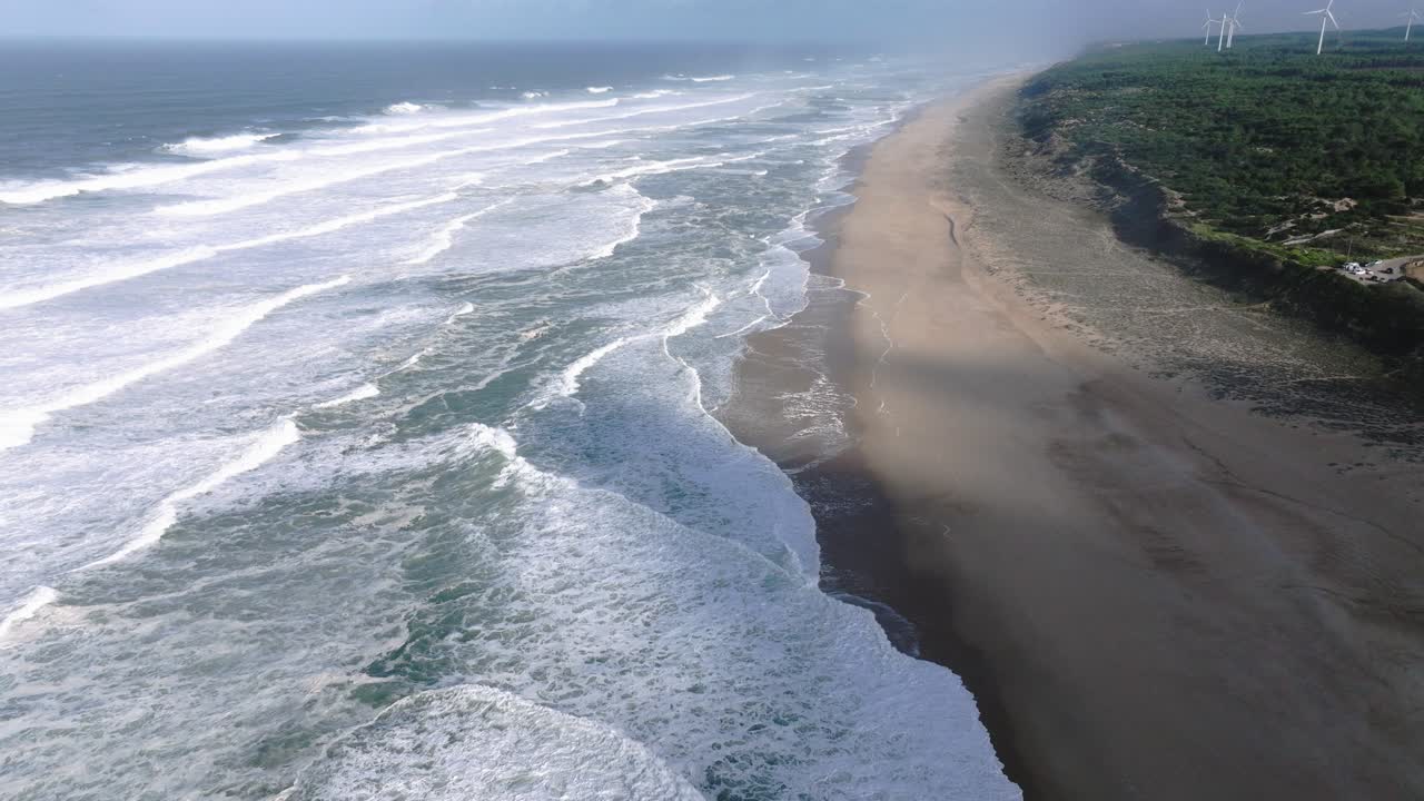 Drone aerial along Nazare Portugal beach, long shoreline with Atlantic waves breaking in parallel lines, cinematic coastal seascape with sand and surf