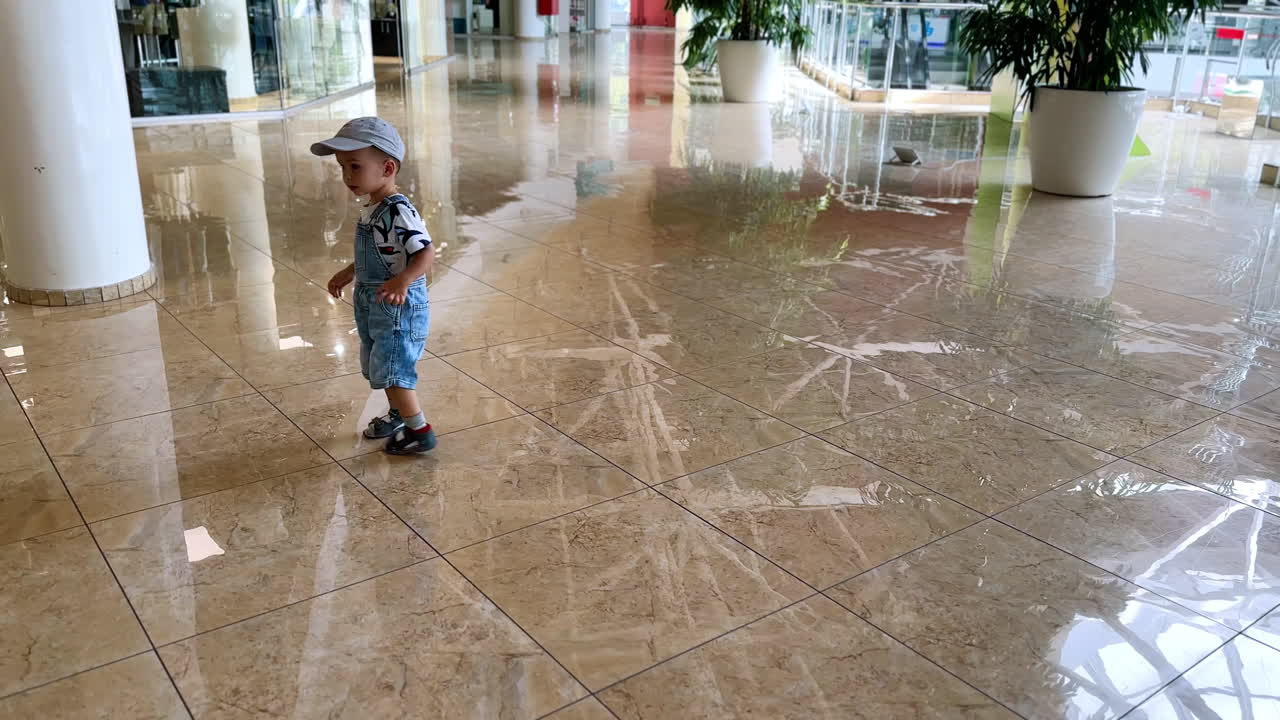 Sweet little boy in a cap is walking by the shiny tiled floor in the mall. Cute kid looking around with interest.