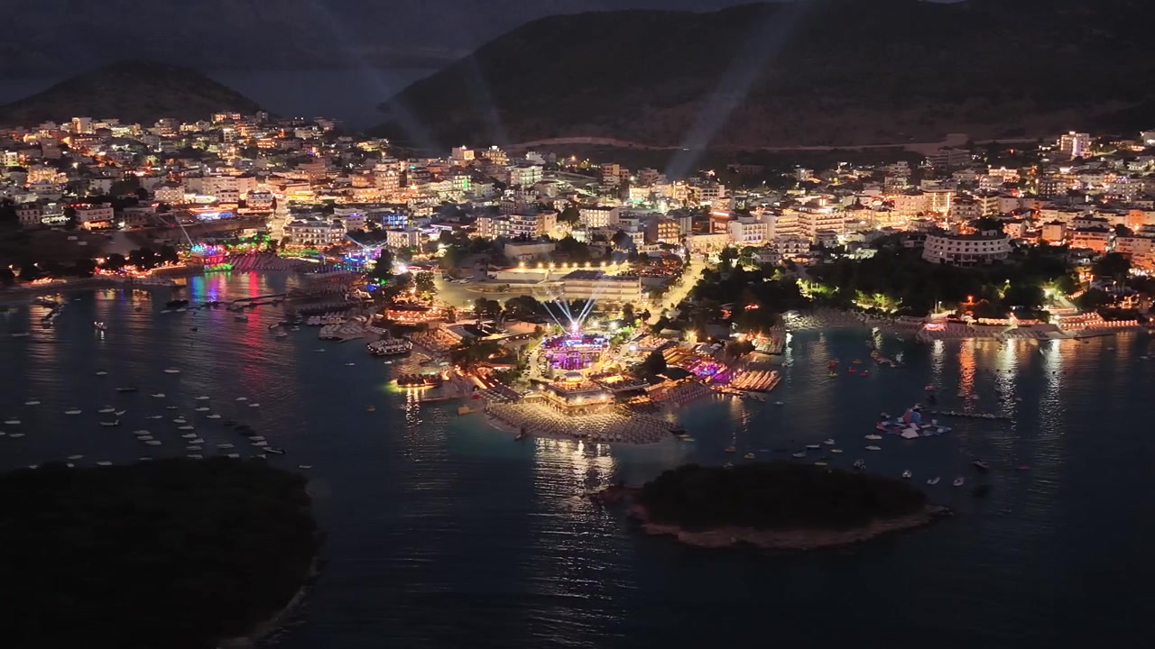 Spettacolare scenario notturno di Ksamil, Albania, catturato da drone. Il filmato evidenzia le strade illuminate della città, i vivaci resort sulla spiaggia e la sagoma scura delle famose isole di Ksamil nella baia