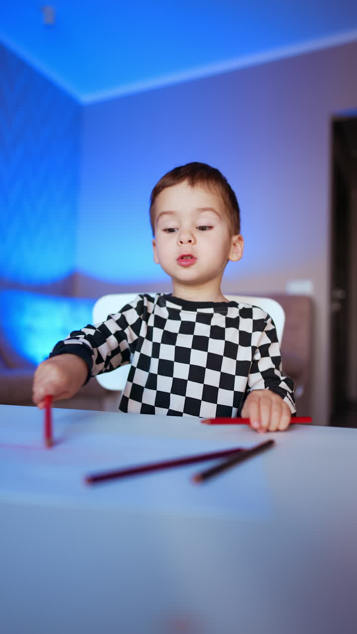 Beautiful Caucasian toddler boy sits at desk. Cute kid moves the red pencil by the paper. Baby boy drawing at home. Vertical video.