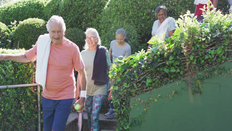 un grupo diverso de adultos felices de sexo masculino y femenino que llegan a una soleada cancha de tenis al aire libre, en cámara lenta