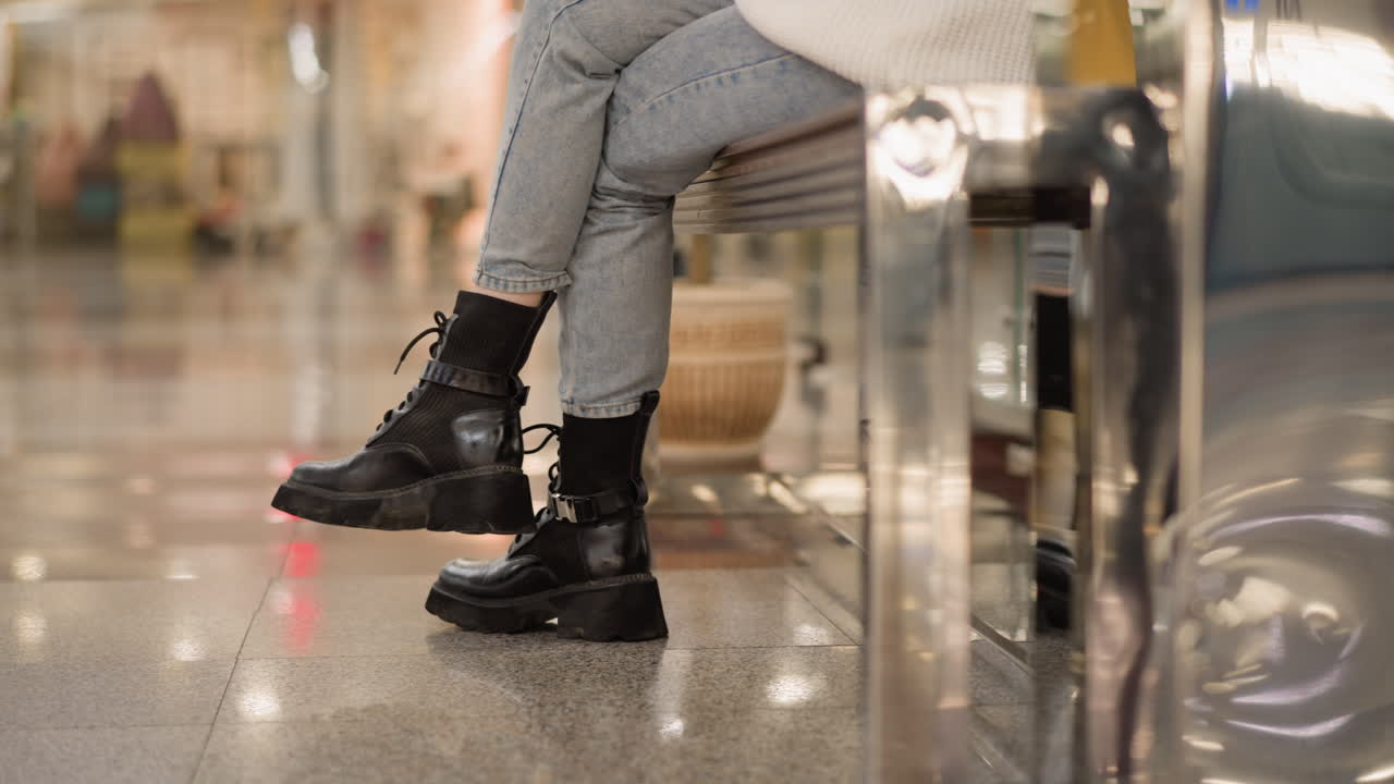 cross leg of trendsetter seated on mall bench wearing chunky black boots and denim jeans showcasing casual fashion detail inside bright shopping mall under ceiling lights captured from side angle
