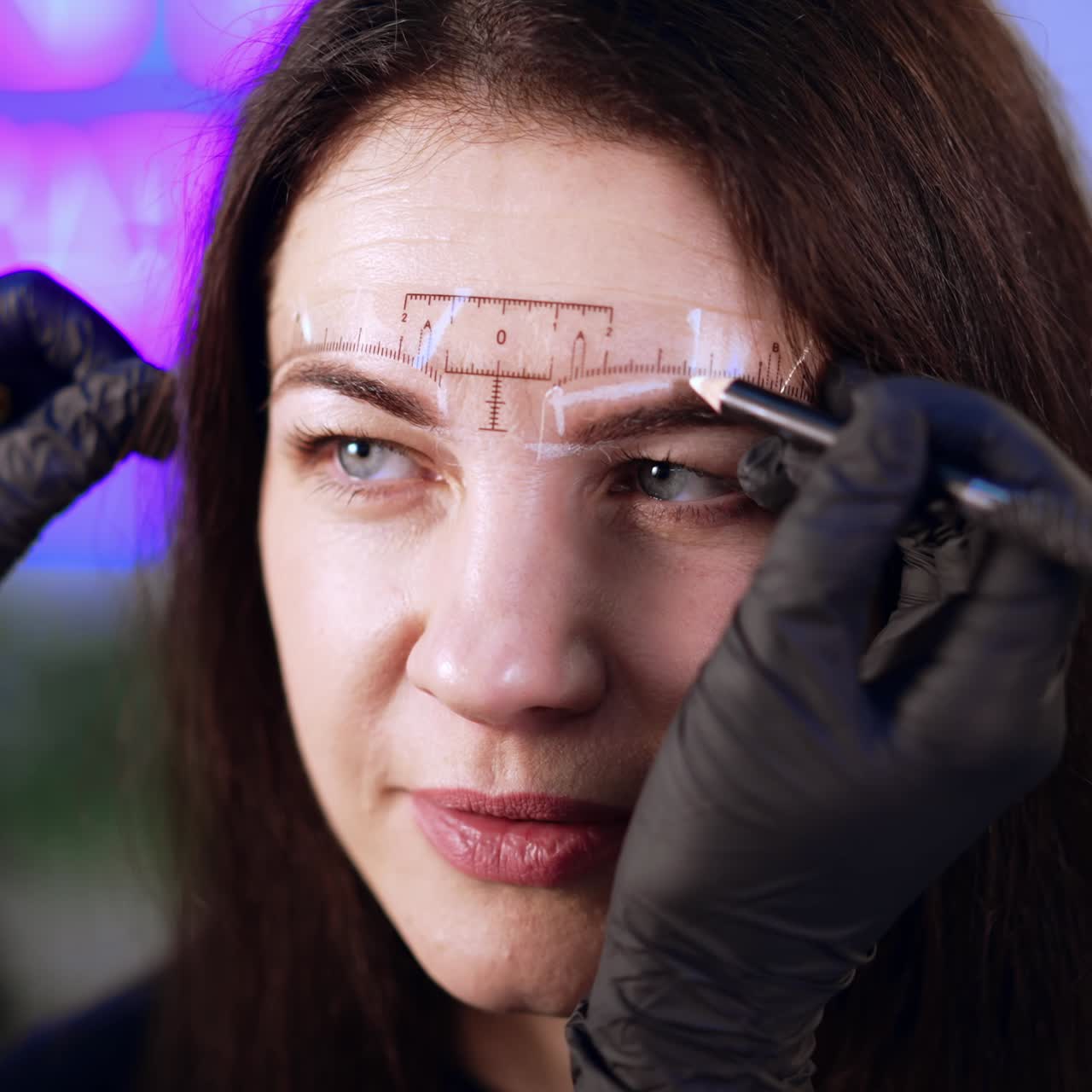 Black gloved hands of beautician uses white pen to mark eyebrow lines for the client. Brunette woman is prepared for eyebrow permanent make up