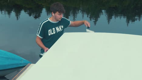 young man finishes thinned coat of white primer on wooden boat roof