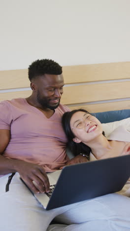 vertical video of happy diverse couple lying and using laptop in bedroom