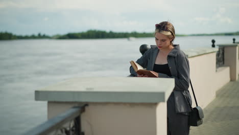 woman in grey clothing rests her right hand on an iron fence while reading a book by the riverfront, the wind softly blows the book s pages, with distant trees and water visible in the background