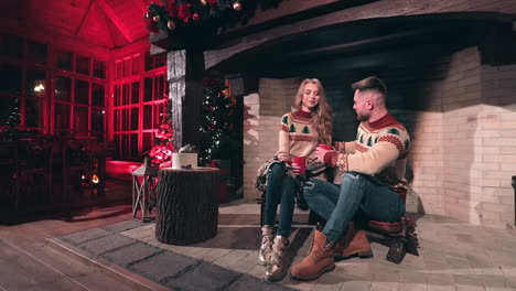 romantic couple in knitted christmas sweaters. young man and woman sitting near the fireplace and drinking tea on christmas decorated room background.