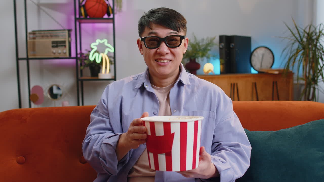 Asian man sits on couch eating popcorn and watching interesting tv serial sport game online at home