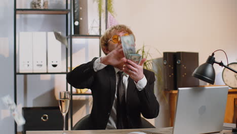 Cheerful-rich-business-man-working-on-office-laptop-wasting-throwing-money-to-camera-profit-win