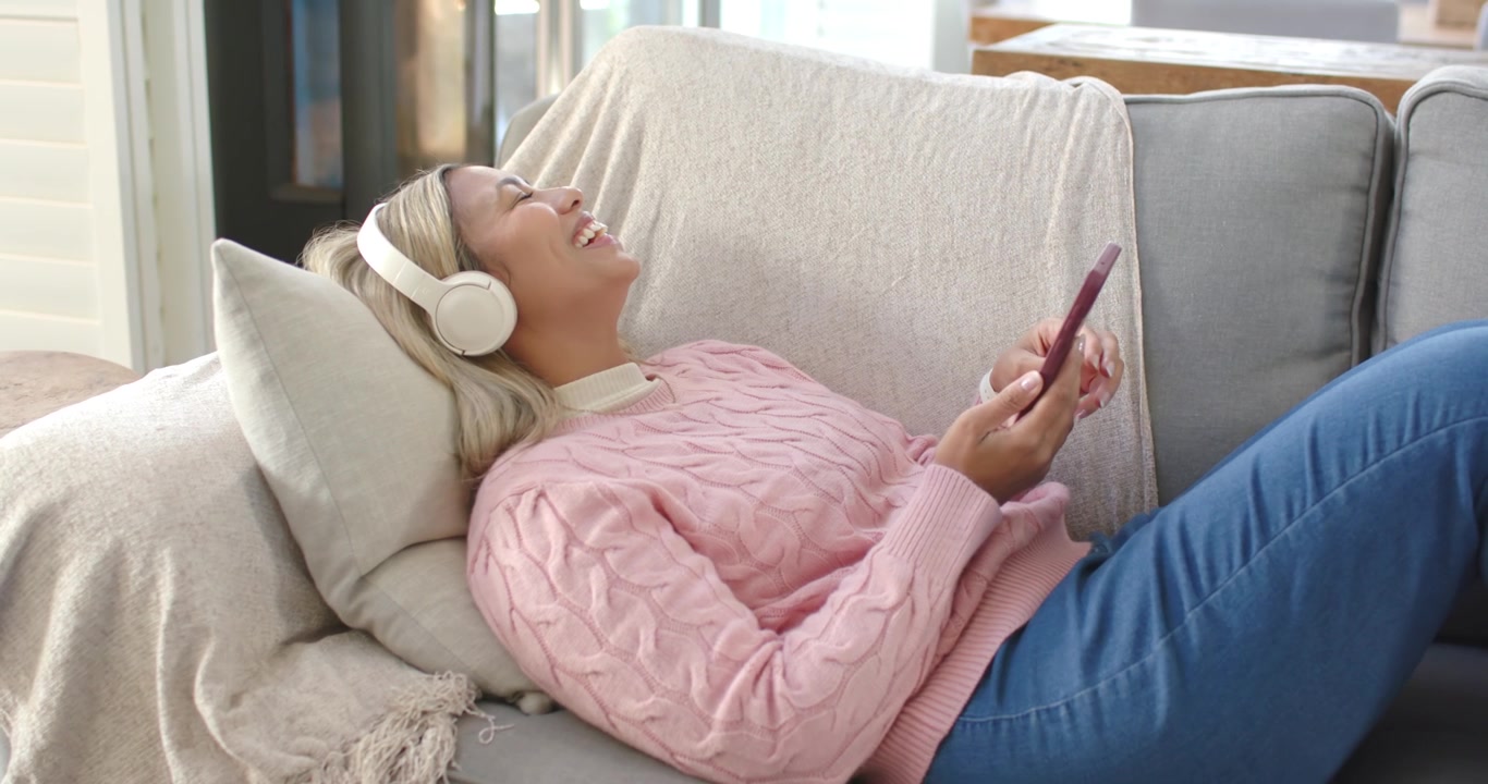 Donna afroamericana che riposa sul divano con cuffie a maglia rosa, telefono che vibra, che tocca e ride, con spazio per il testo.