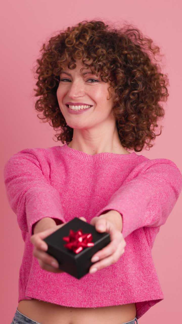 Cheerful young woman giving gift box in pink studio. Vertical footage