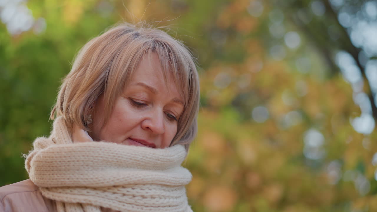 close up of middle aged woman standing in autumn forest, thoughtful expression as she focuses intently downward, soft scarf around neck and gentle bokeh foliage backdrop