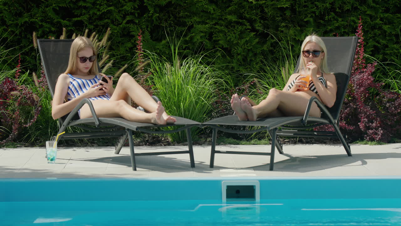 A woman and her daughter are relaxing on sun loungers by the pool, drinking soft drinks