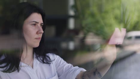 mujer inteligente concentrada pegando notas adhesivas en el vidrio de la ventana planeando negocios en el interior. retrato de ocupado centrado gerente caucásico inteligente pensando de pie en la oficina. cámara lenta