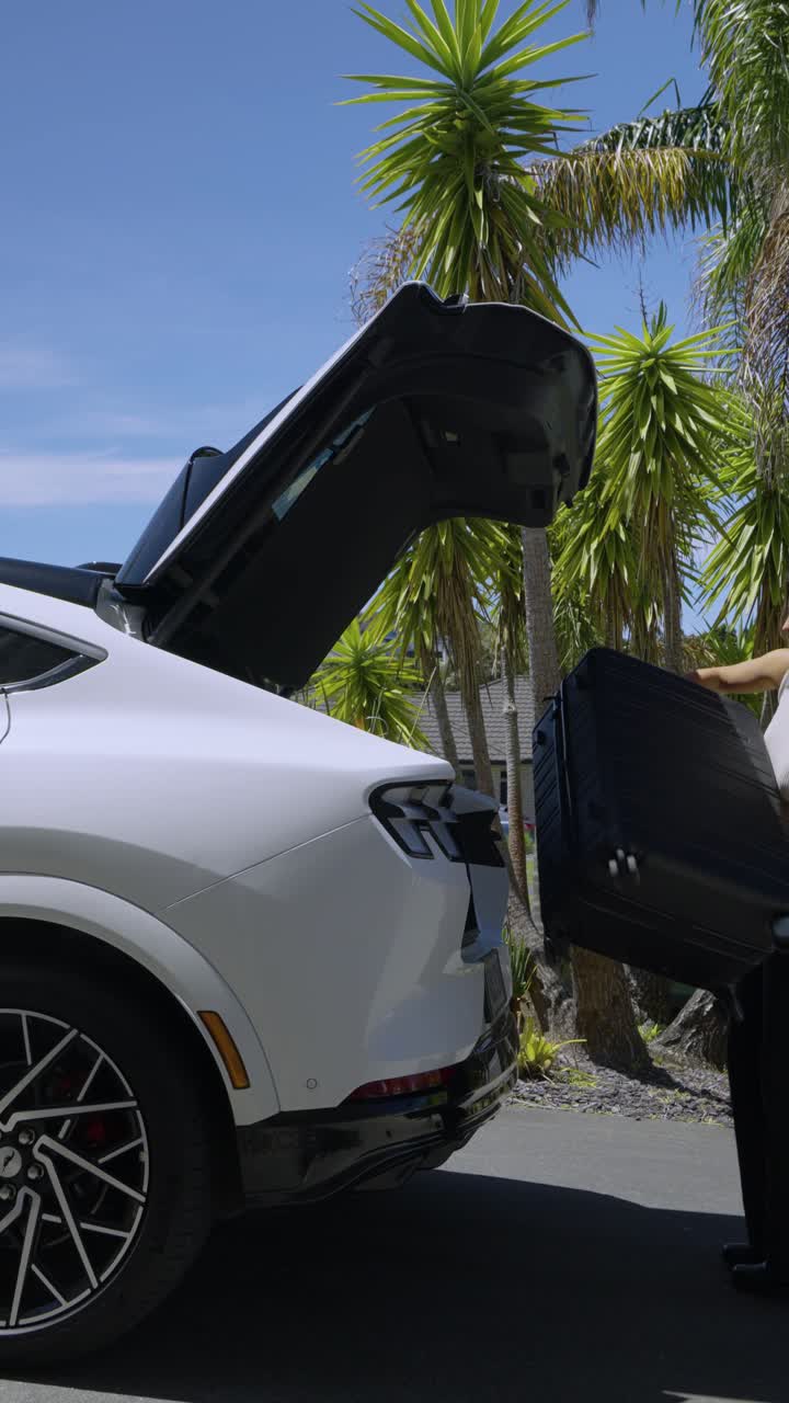 A smartly dressed woman lifts a suitcase and places it into the trunk of a car, preparing for travel.
