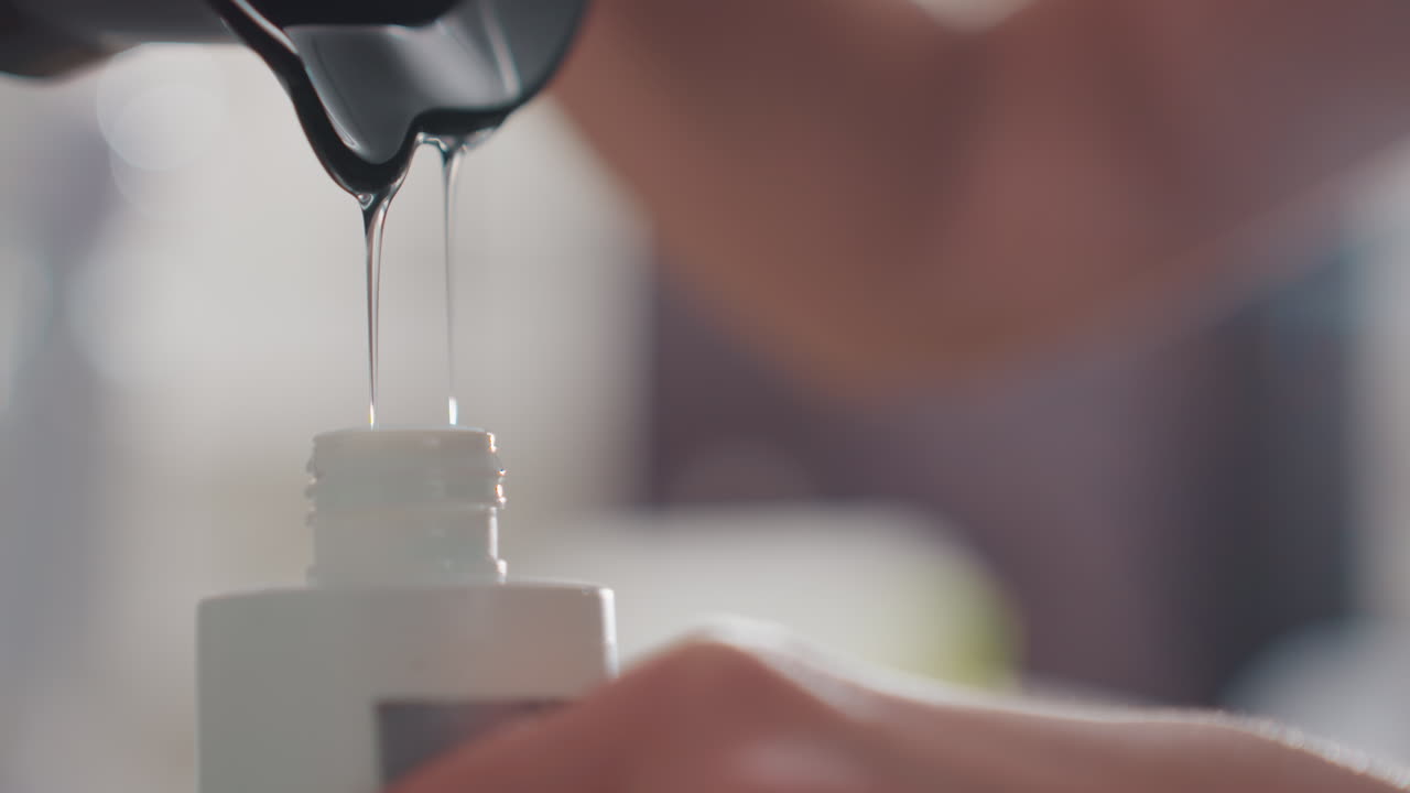 Primer plano de una botella rellenándose desde una jarra, un colorista caucásico vierte un concentrado transparente en una botella pequeña, con manos firmes, retroiluminación brillante, medición cuidadosa y goteo lento, escena de preparación en un salón.