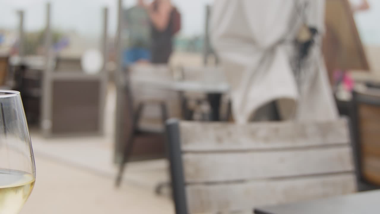 Slow camera pan reveals white wine glasses on outdoor wooden table, beachside cafe, natural daylight
